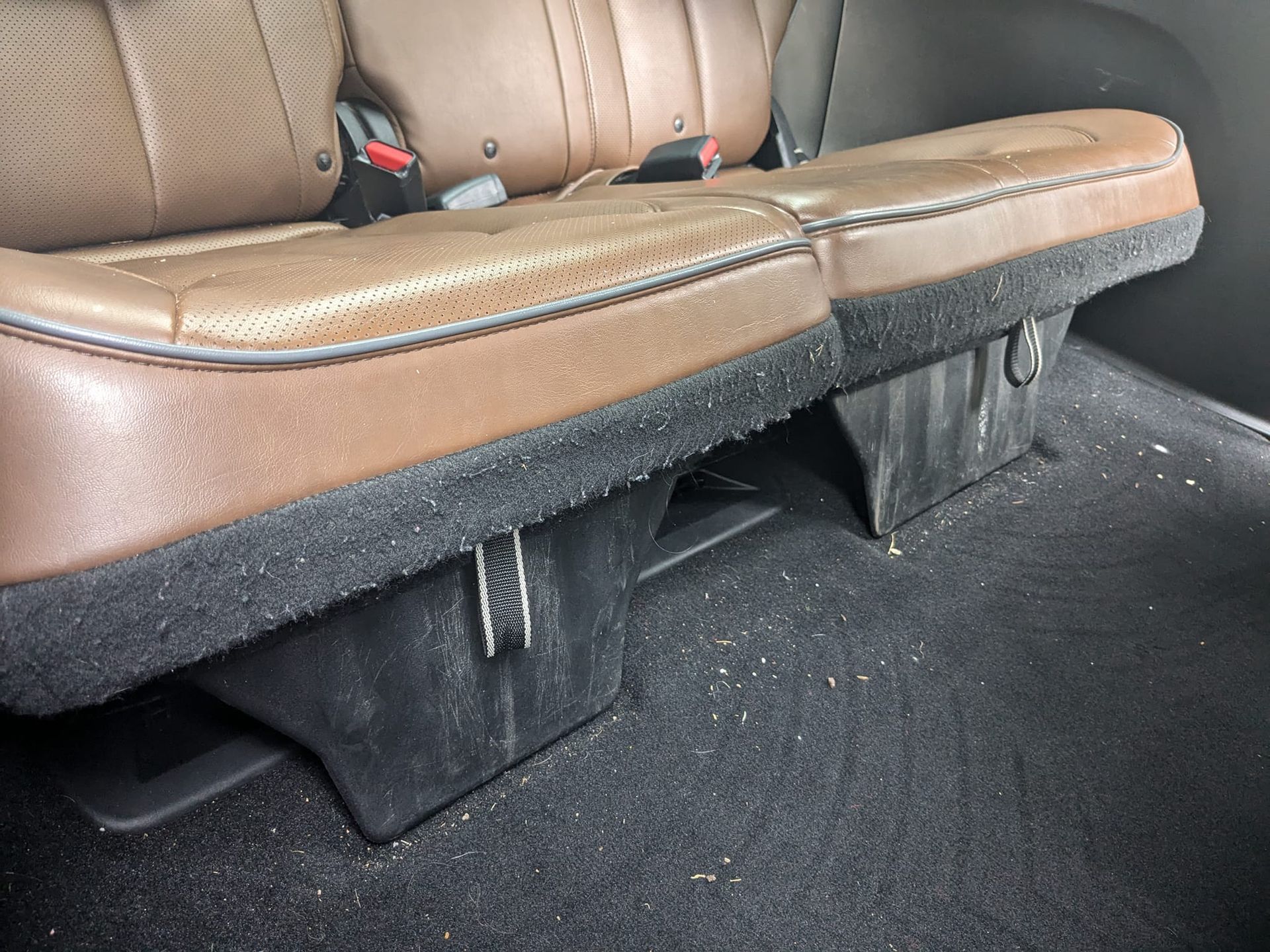 A close-up view of tan leather vehicle seats above black plastic storage compartments and dark carpeted flooring.