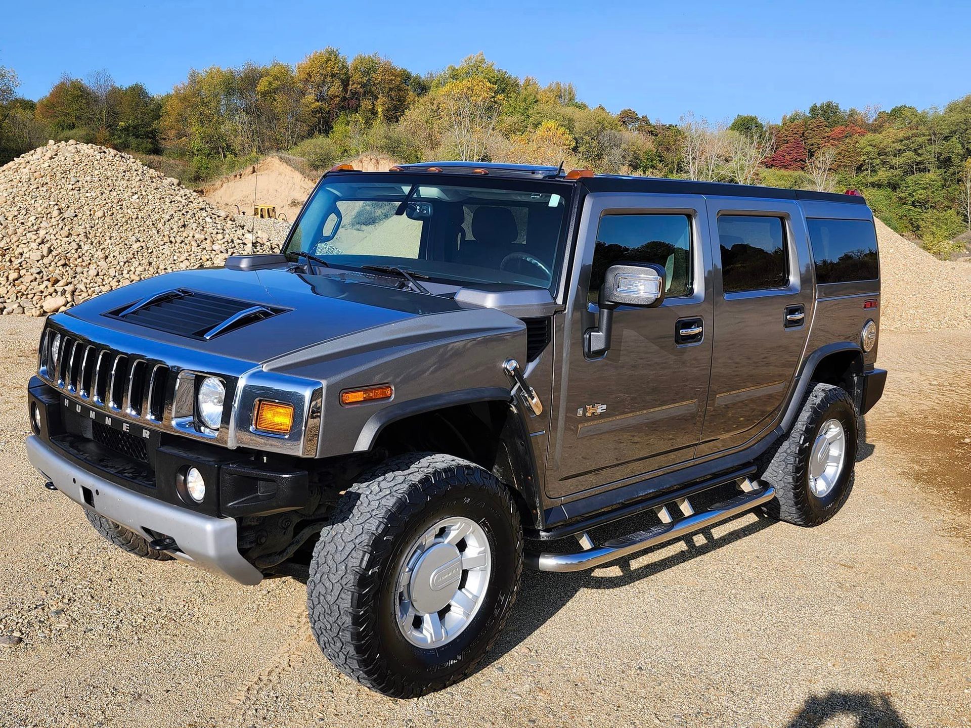 A grey Hummer H2 parked on a gravel surface with a backdrop of trees and a pile of rocks under a clear blue sky.