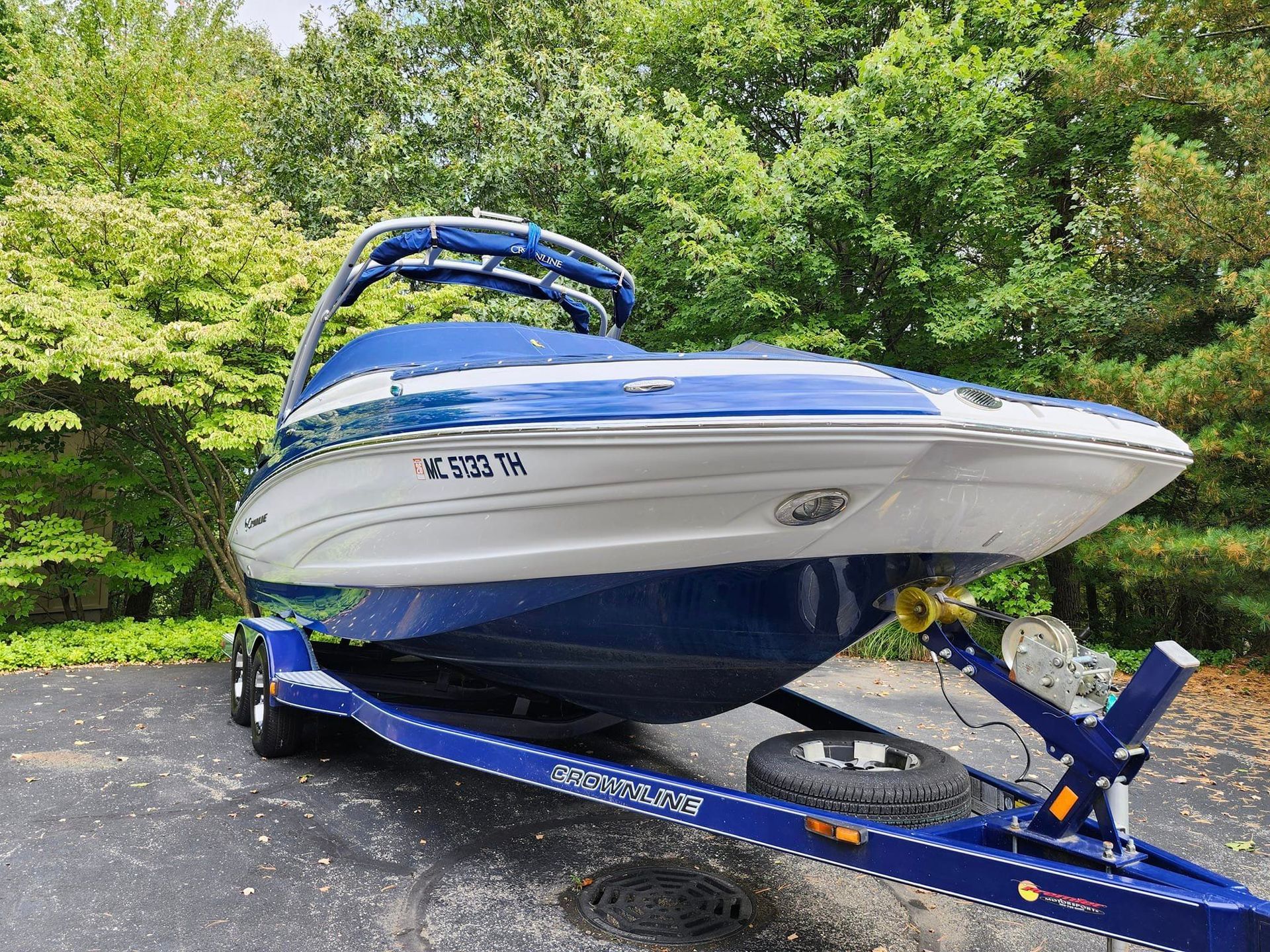 A blue and white motorboat secured on a blue trailer, parked on a paved driveway surrounded by trees.