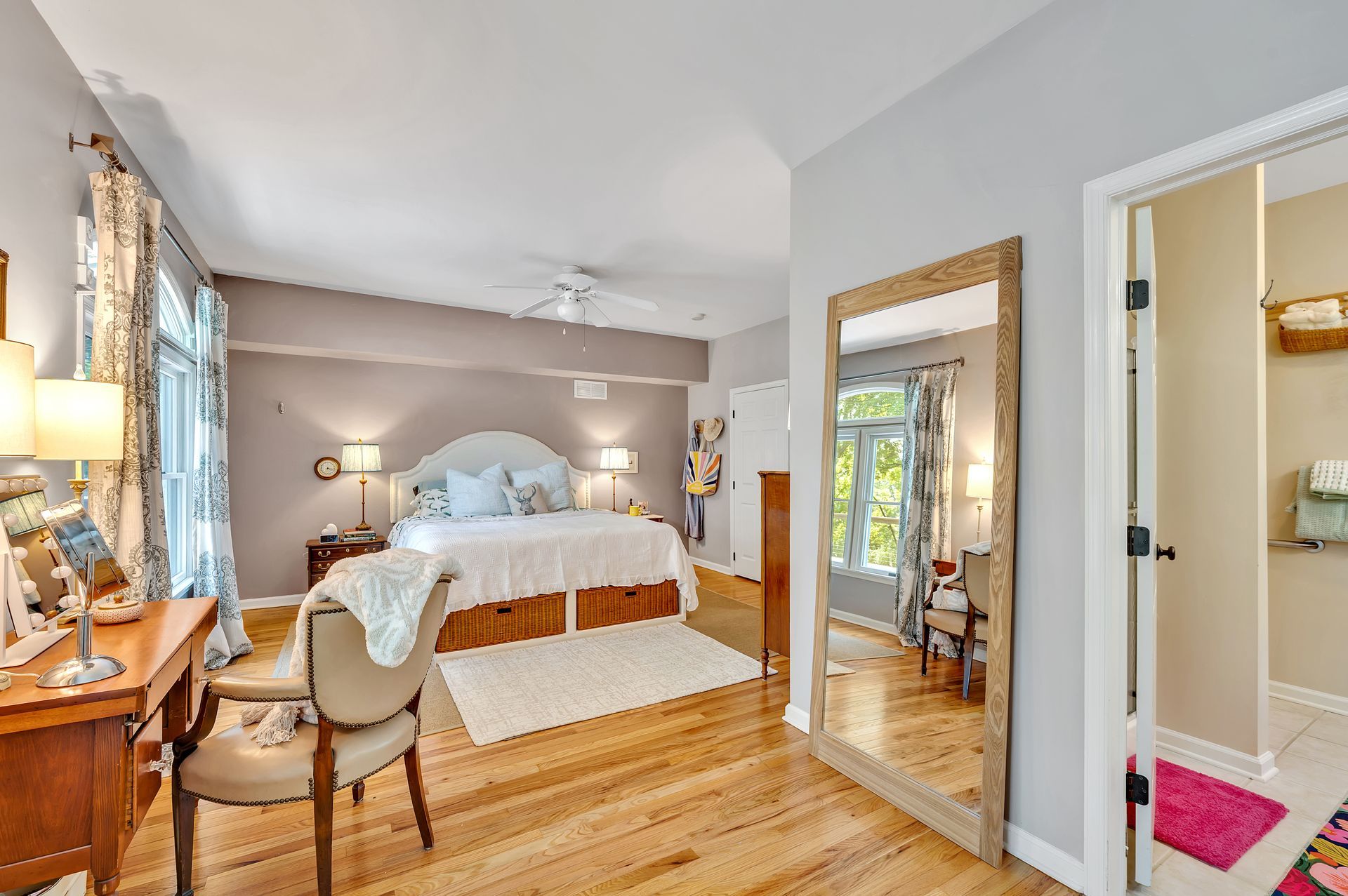 A bright bedroom with a queen bed, hardwood floors, a wooden desk with a chair, and a large floor-length mirror.