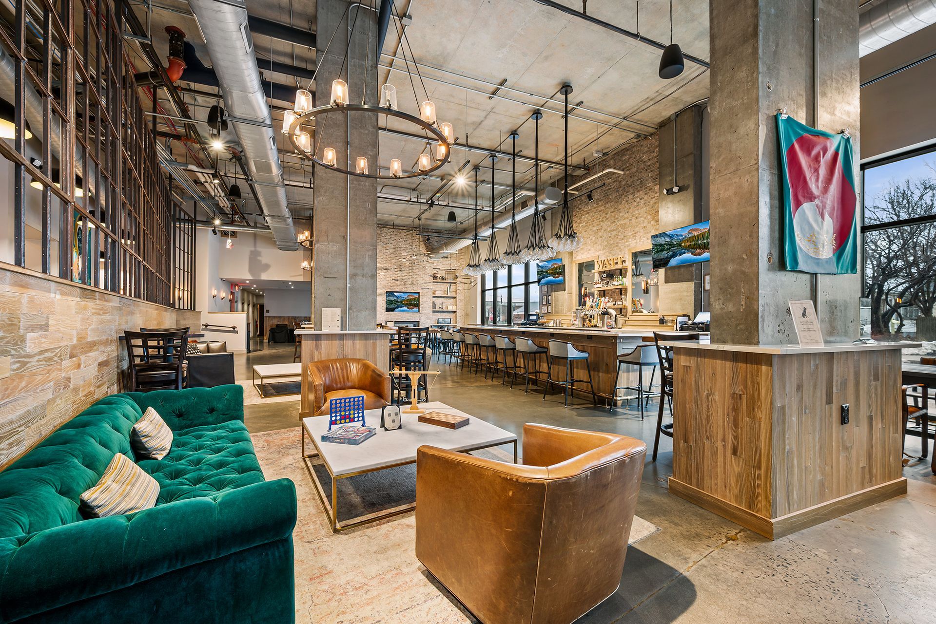 A trendy, open-concept bar lounge with a green velvet sofa, leather armchairs, exposed concrete pillars, and a long bar.