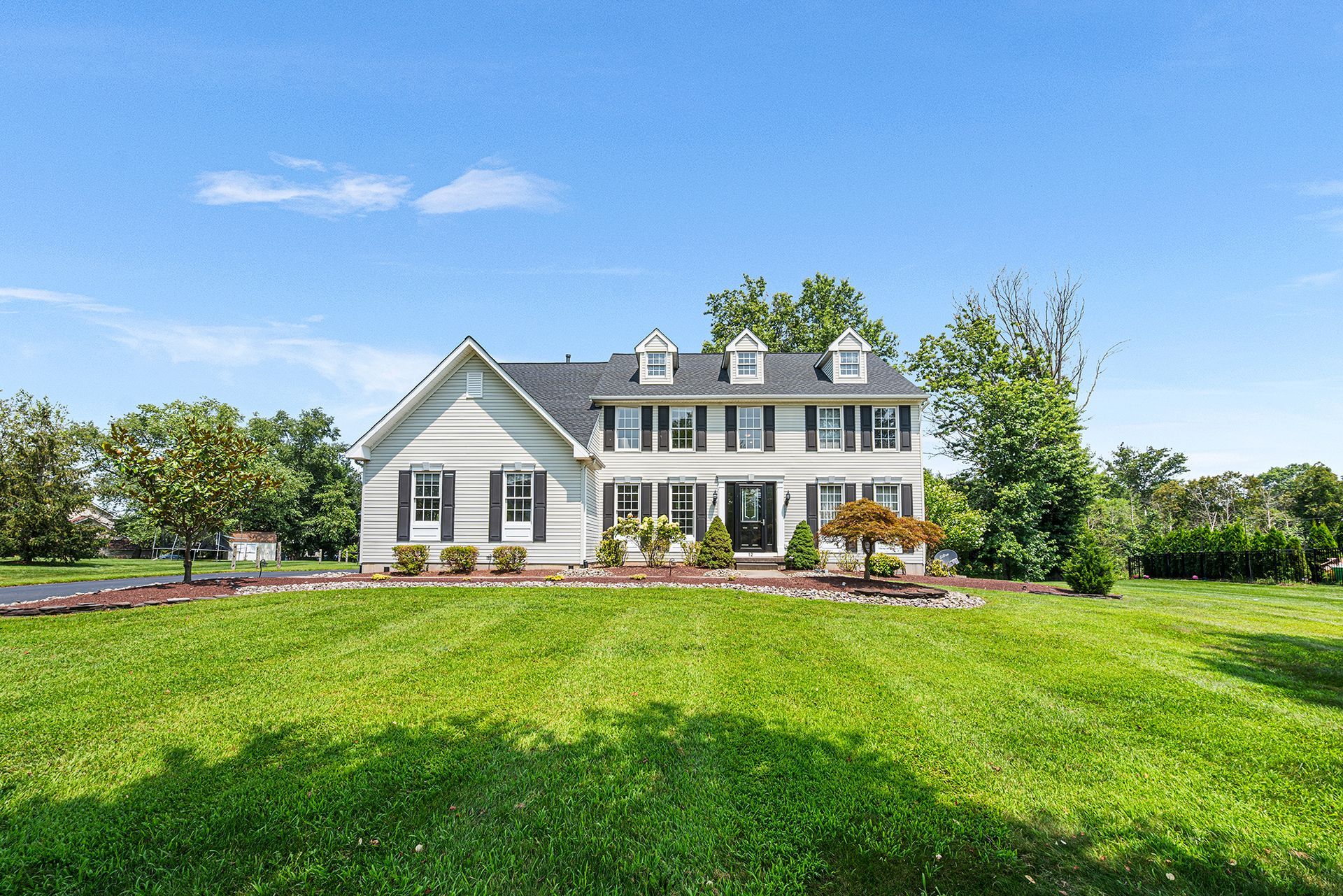 Two-story white house with a dark roof and black shutters, situated behind a wide green lawn under a bright blue sky.