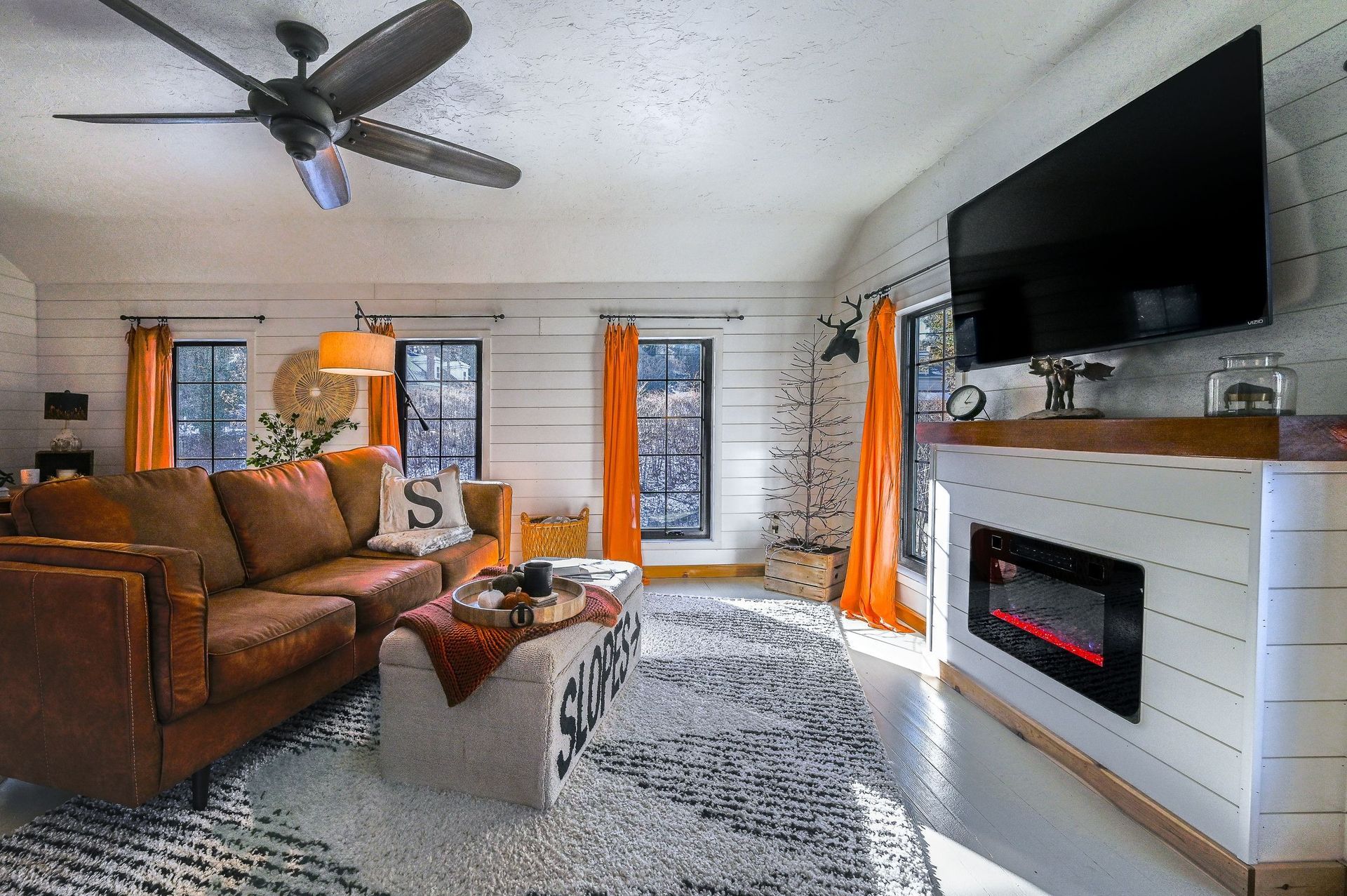 A rustic living room with a brown leather sofa, white shiplap walls, a fireplace with a mounted TV, and orange curtains.