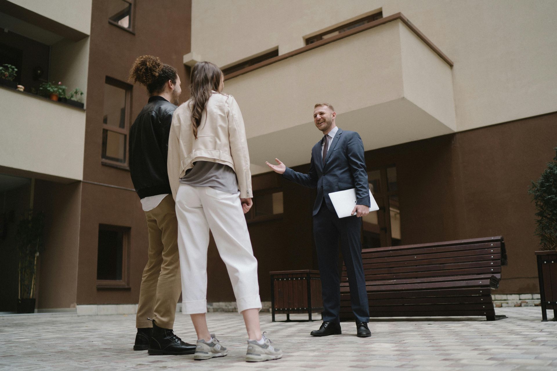 A real estate agent in a suit gesturing towards a building while talking to two people standing in a courtyard.