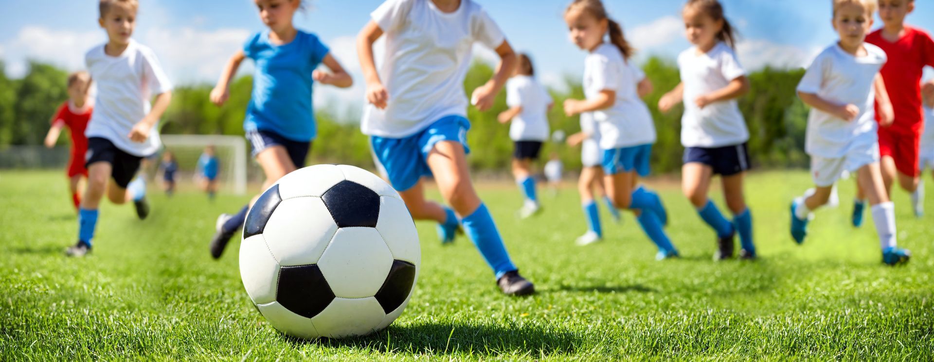 A group of children in sports uniforms running across a grassy field toward a soccer ball.