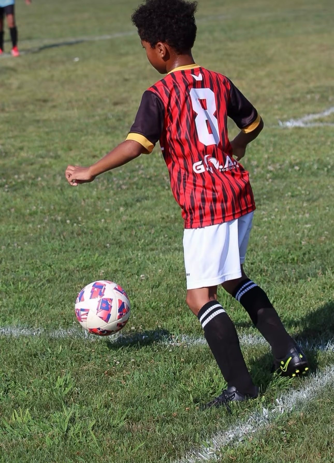 A young soccer player in a red and black striped jersey with the number 8 dribbles a soccer ball on a grassy field.