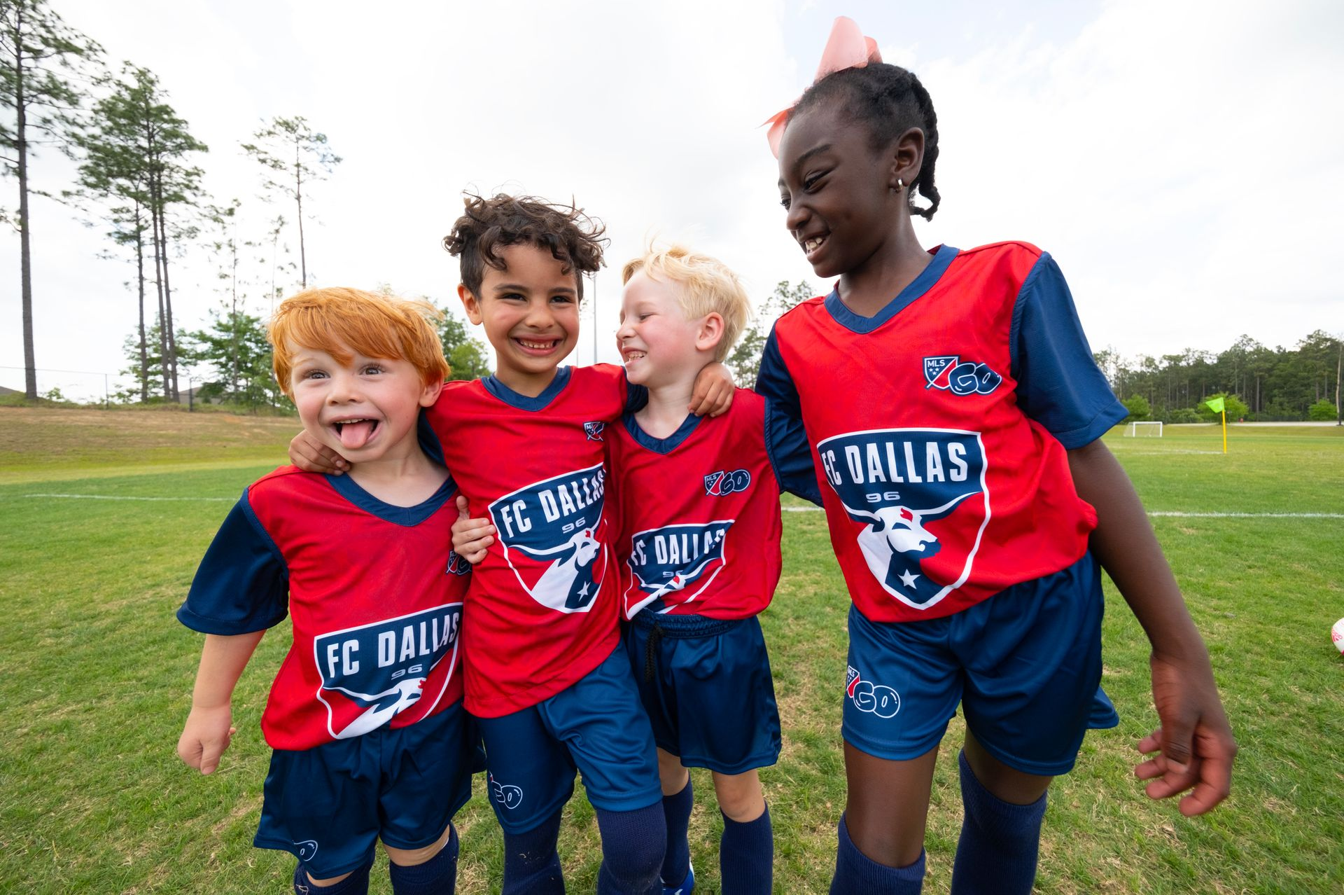 Four children in red and navy FC Dallas soccer uniforms stand together laughing on a grassy field.