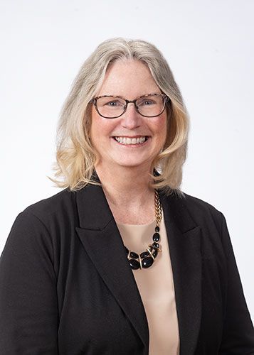 Smiling woman with glasses and shoulder-length blonde hair, wearing a black blazer and necklace on a white background