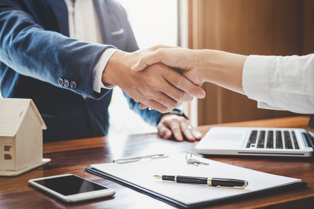 Handshake over a desk with a laptop, notebook, pen, phone, and small house model