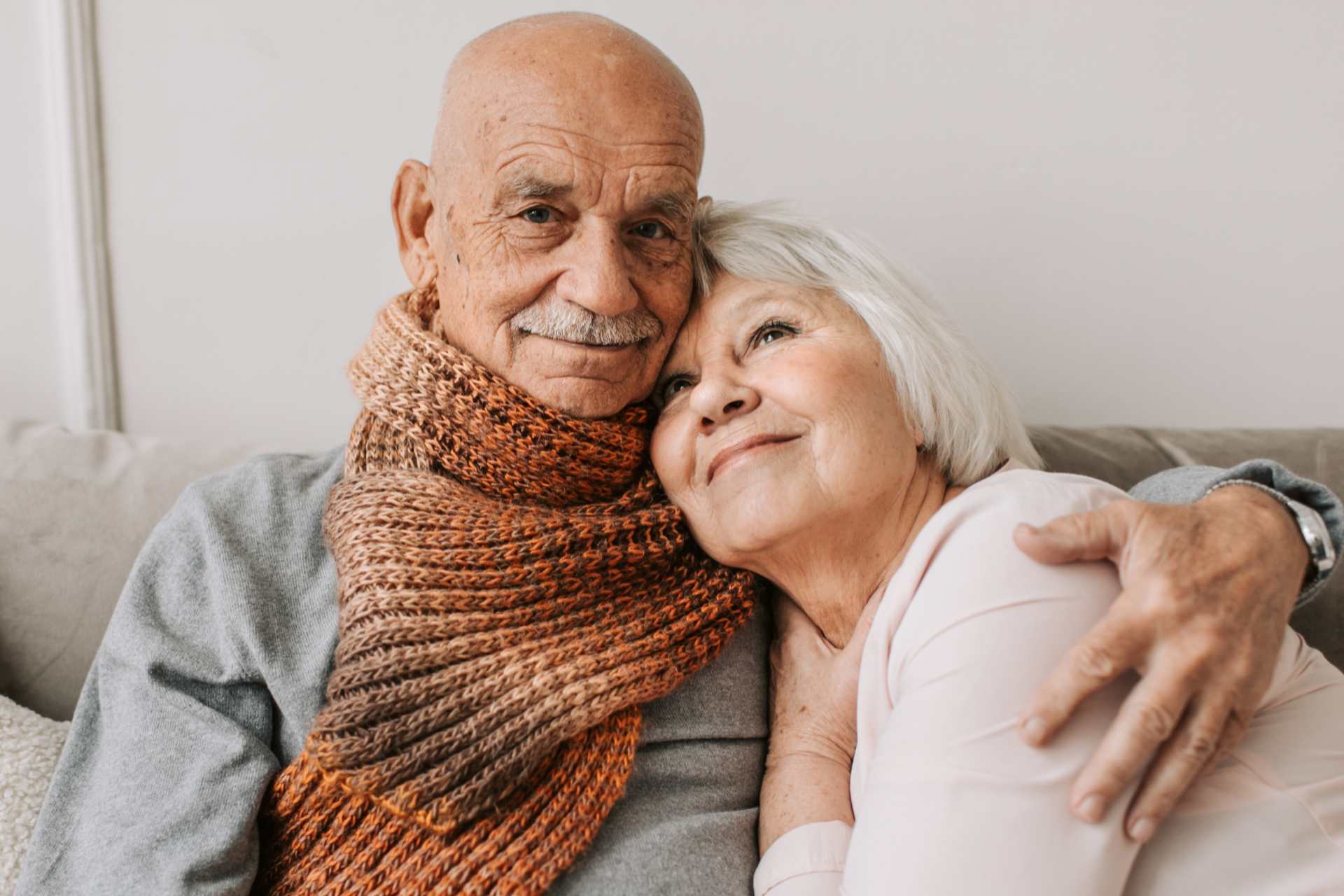 Two elderly people cuddling on a couch, smiling warmly indoors.