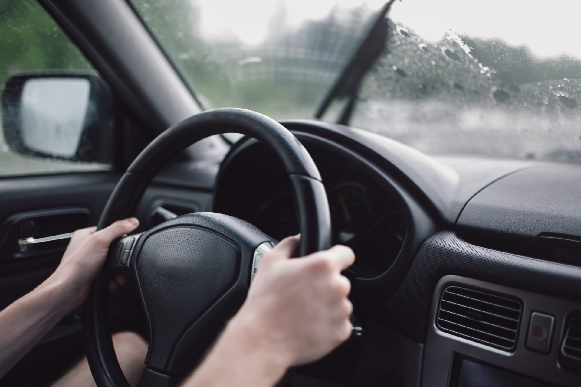 Hands on a steering wheel inside a car on a rainy road.