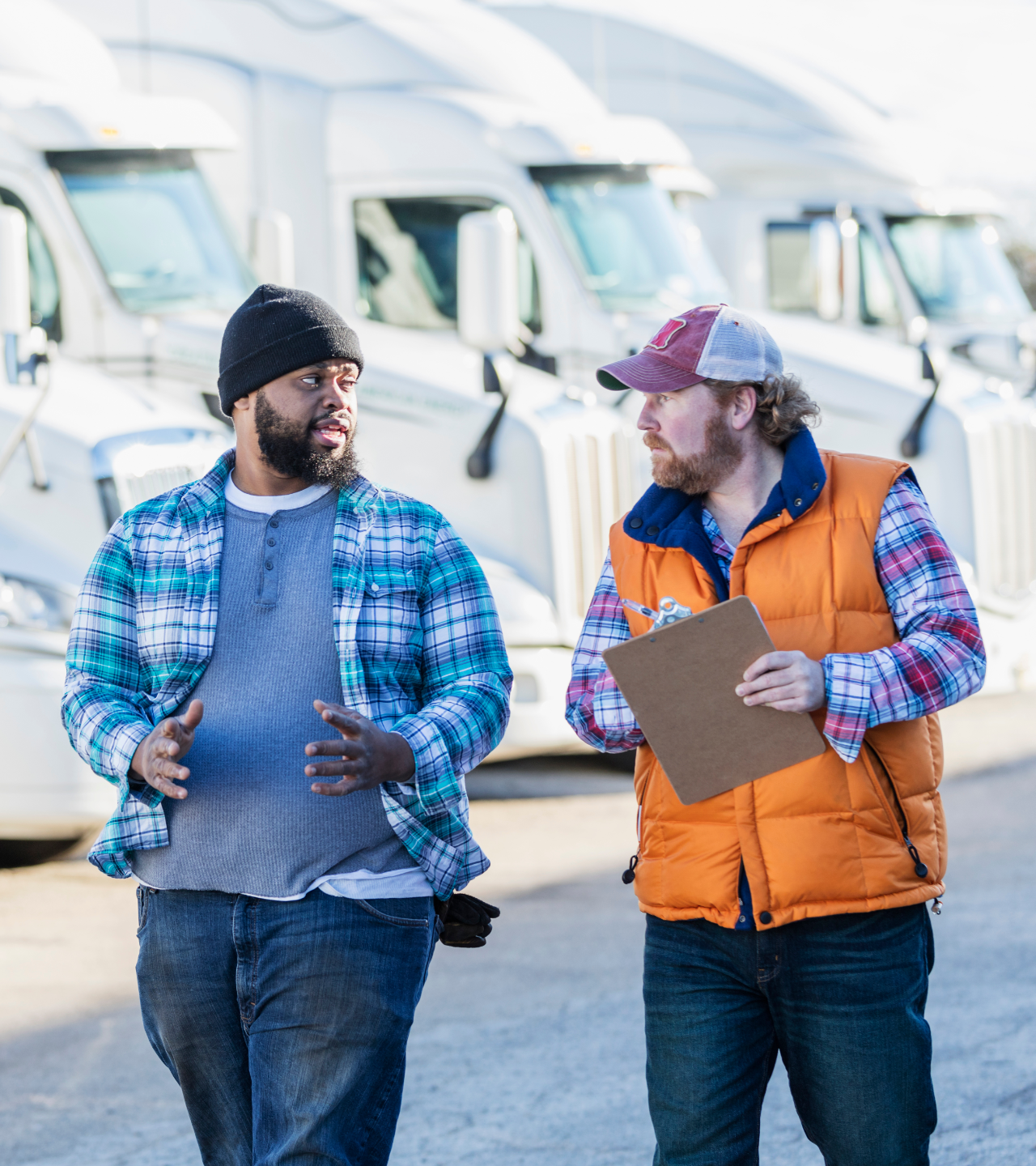 Two workers in winter clothing talk beside parked white semi trucks, one holding a clipboard.
