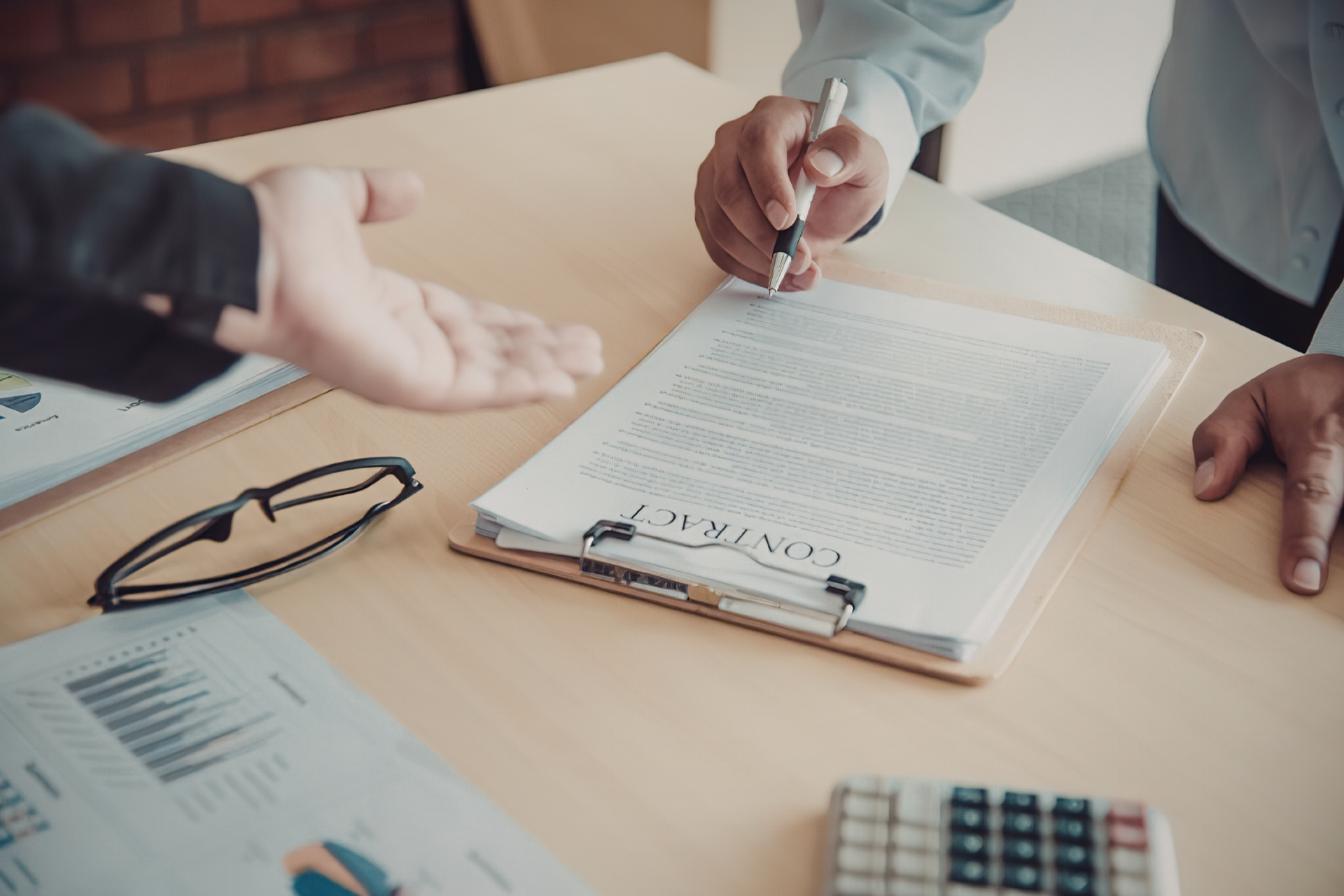 Hands over a clipboard with documents at a meeting table, with glasses and papers nearby.