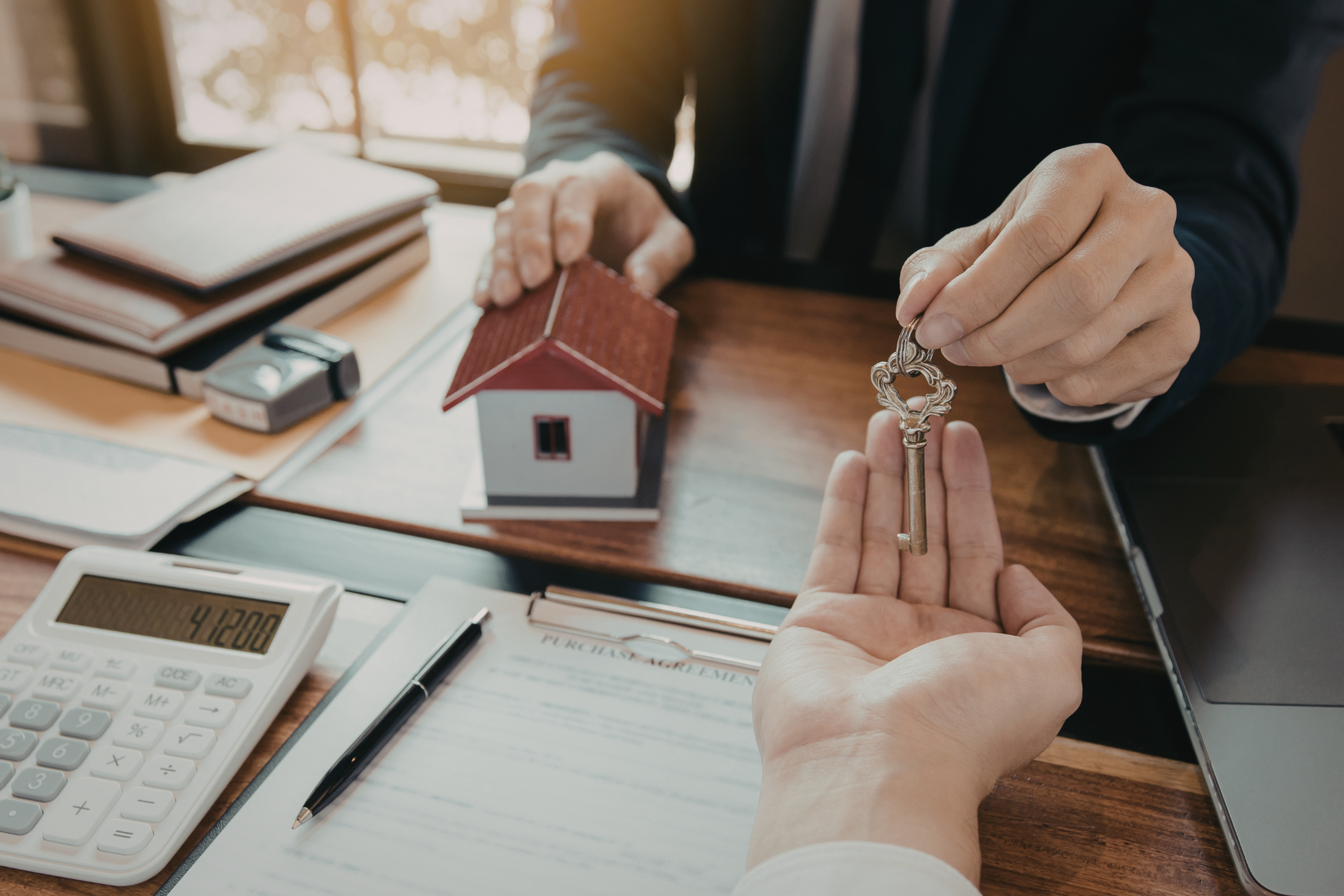 Hands exchanging house keys over a desk with paperwork, calculator, and model house in a real estate office