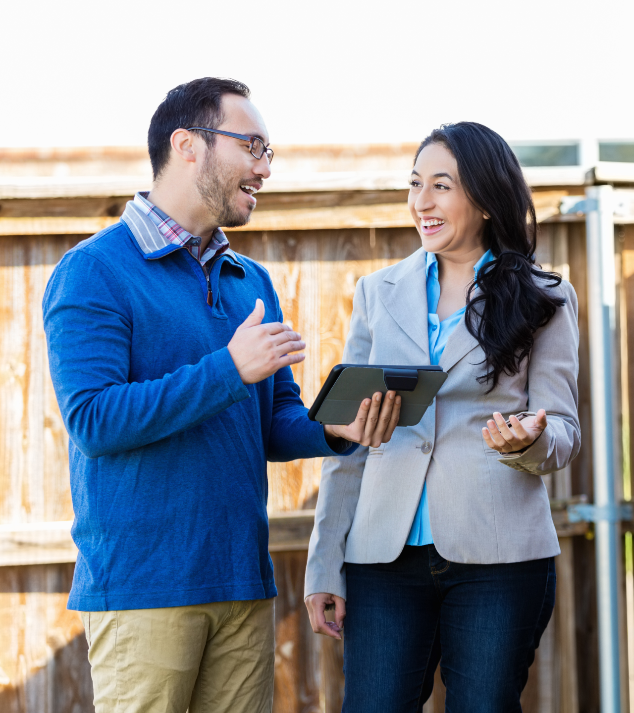 Two people talking outdoors, one holding a tablet and the other a coffee cup, near a wooden fence.