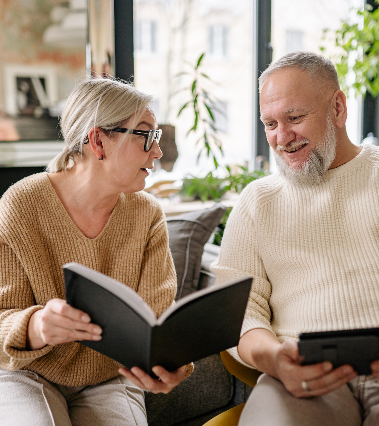 Two people in beige sweaters sit on a couch, smiling and looking at books in a bright living room