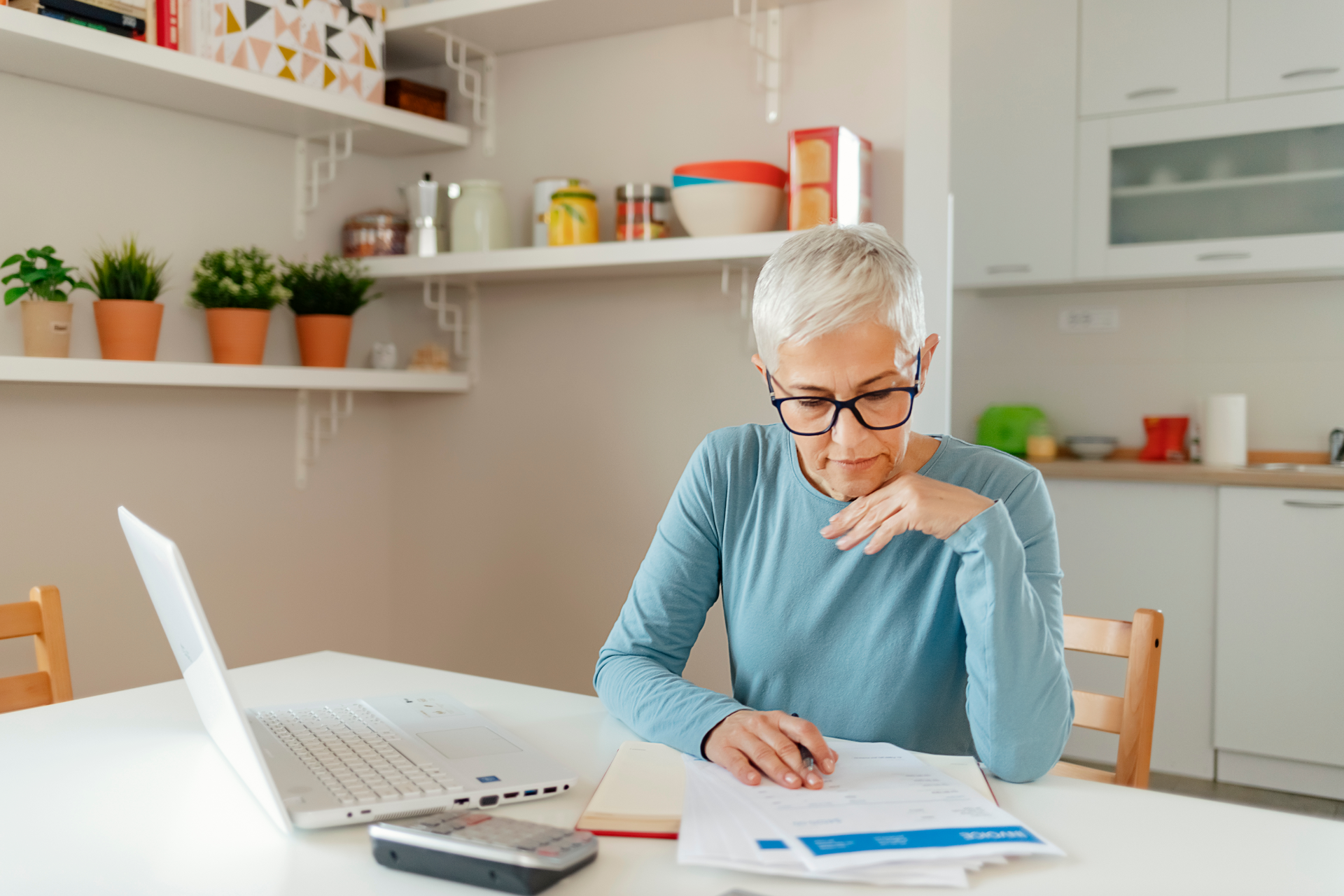 Person writing at a kitchen table beside an open laptop, with plants and shelves in the background