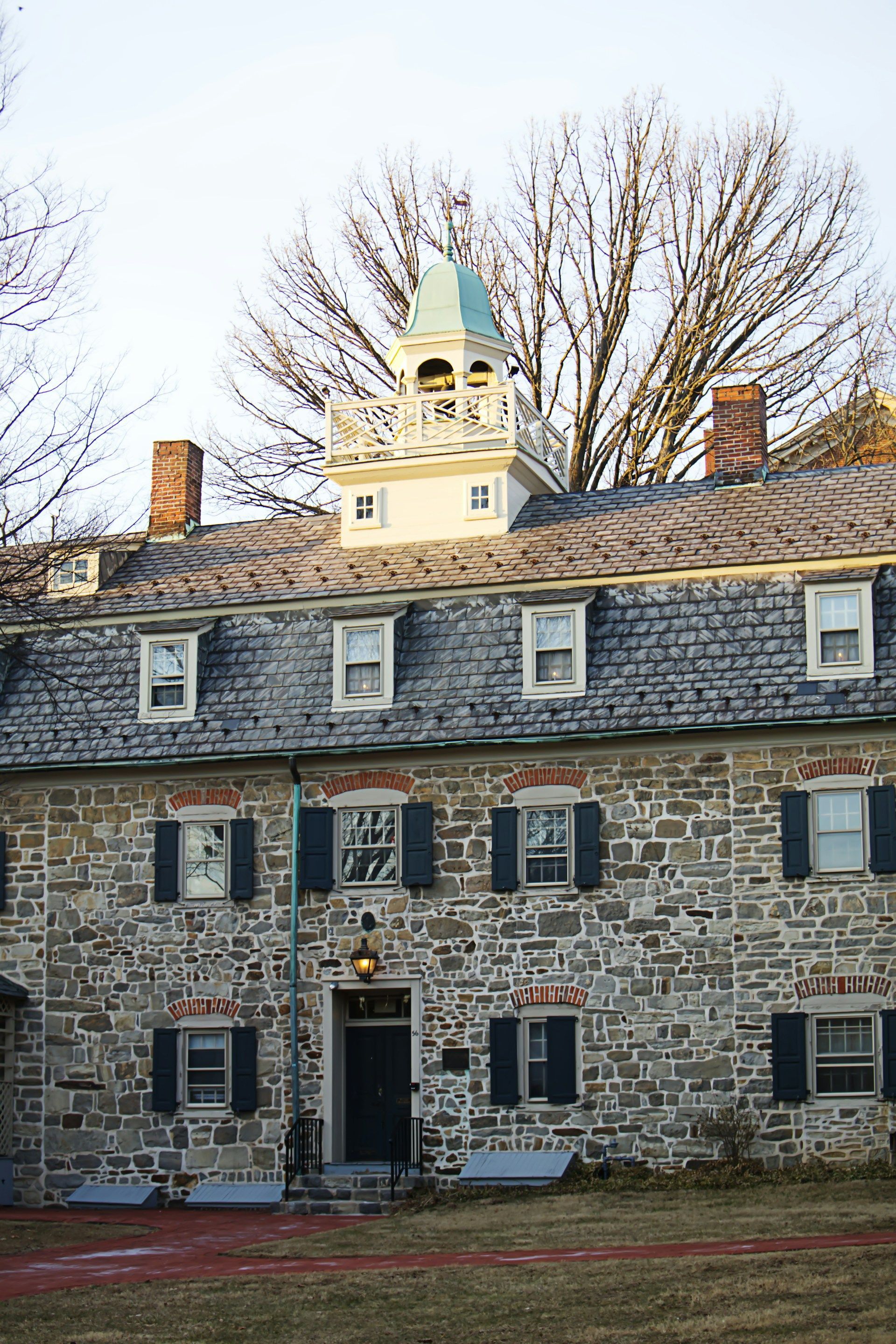 Historic stone building with slate roof, central entrance, and white cupola against bare trees