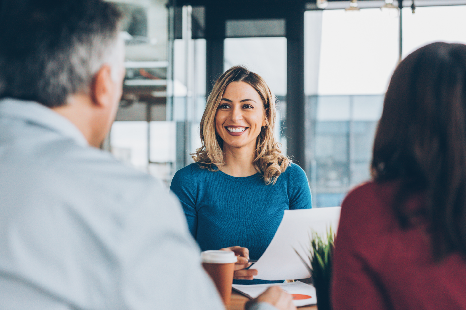Three people in a bright office meeting, with a smiling woman holding a cup at the center.