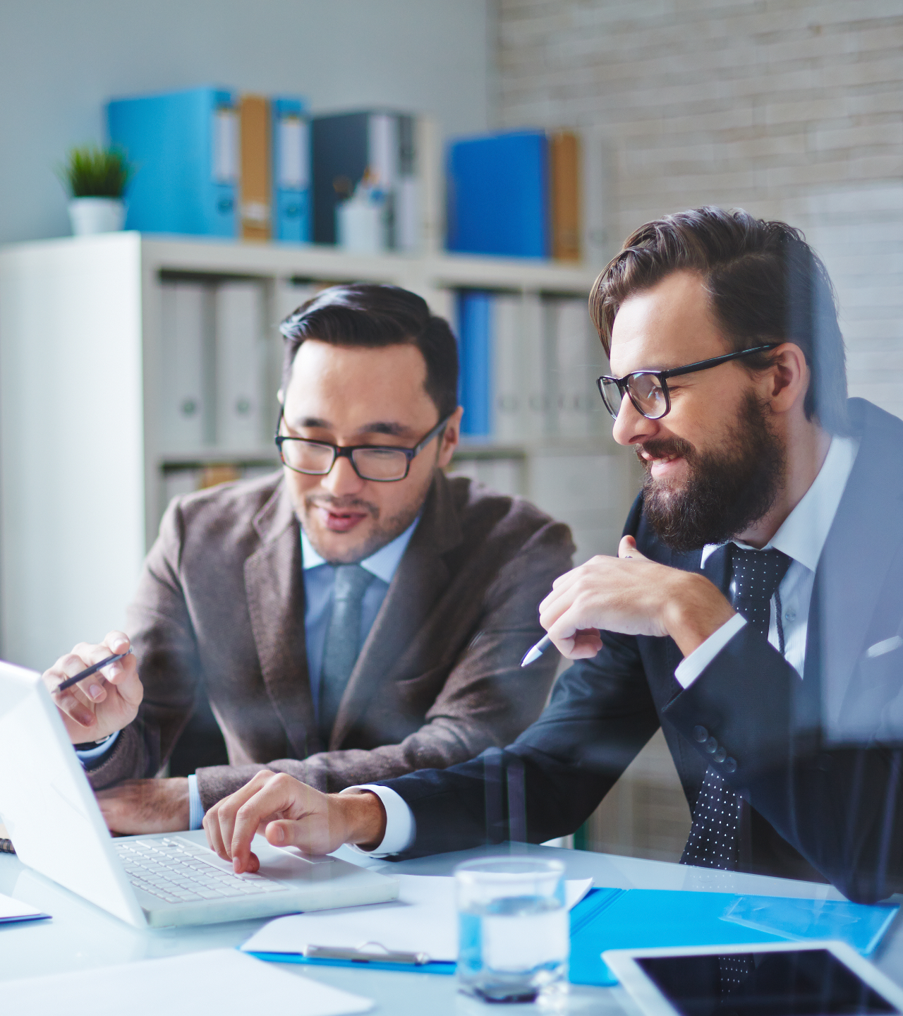 Two coworkers in suits reviewing a laptop and documents in a bright office meeting