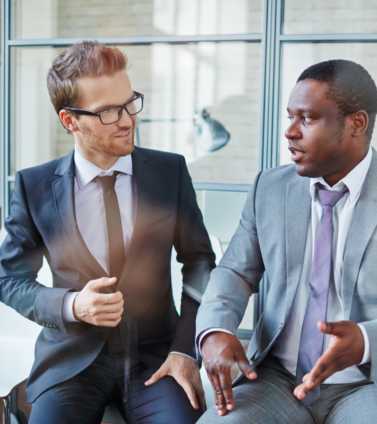 Two businessmen in suits talking on a bench in a bright office lobby