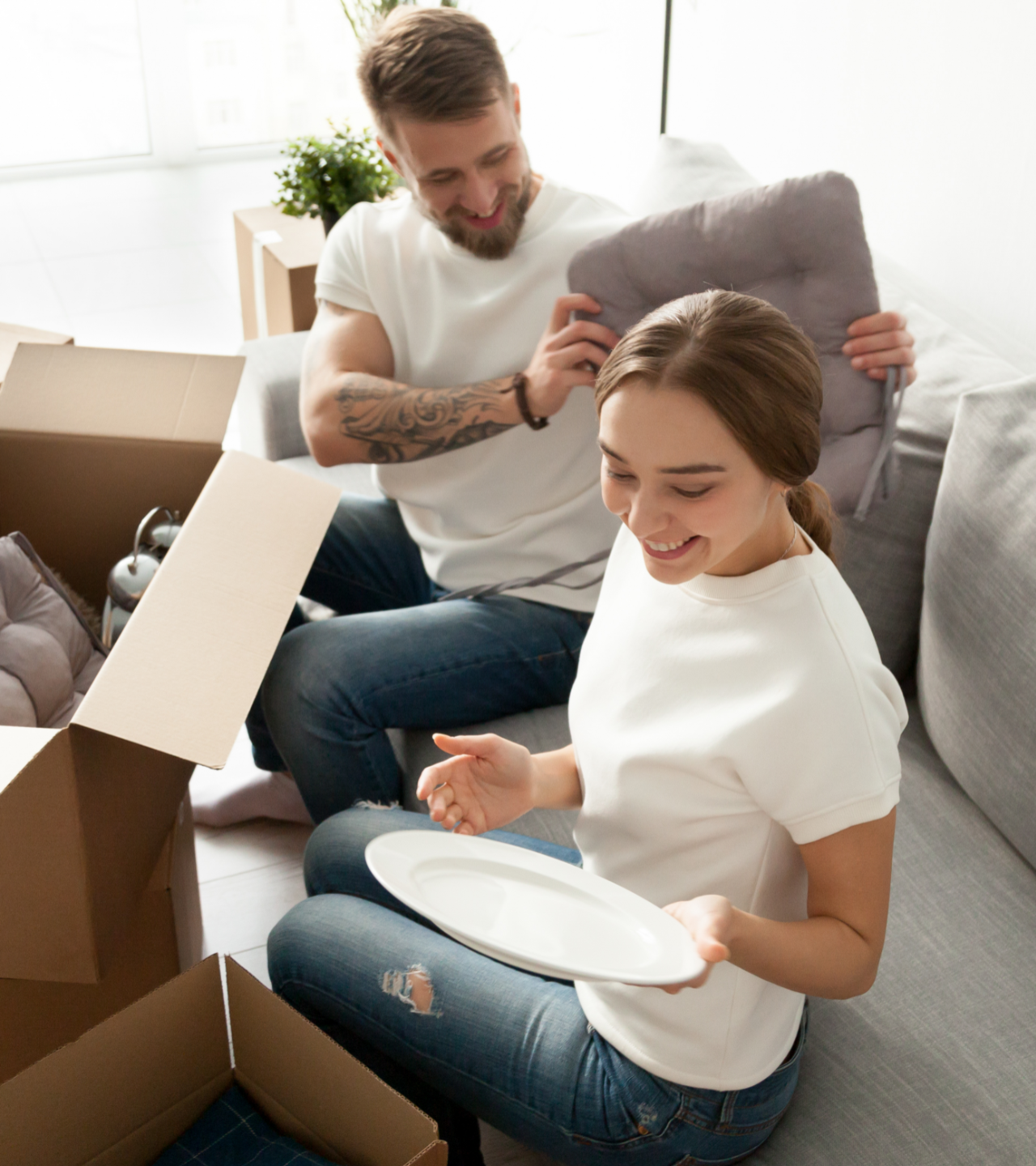 Two people unpacking dishes in a bright living room, smiling beside open boxes and a sofa