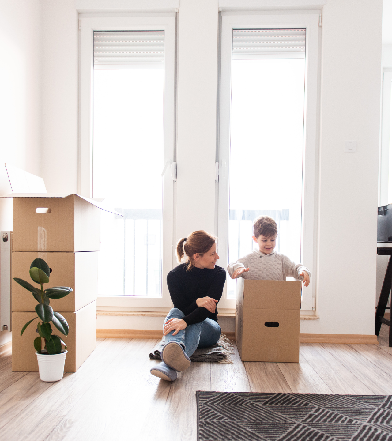 Adult helping child sit in a cardboard box in a bright room with tall windows