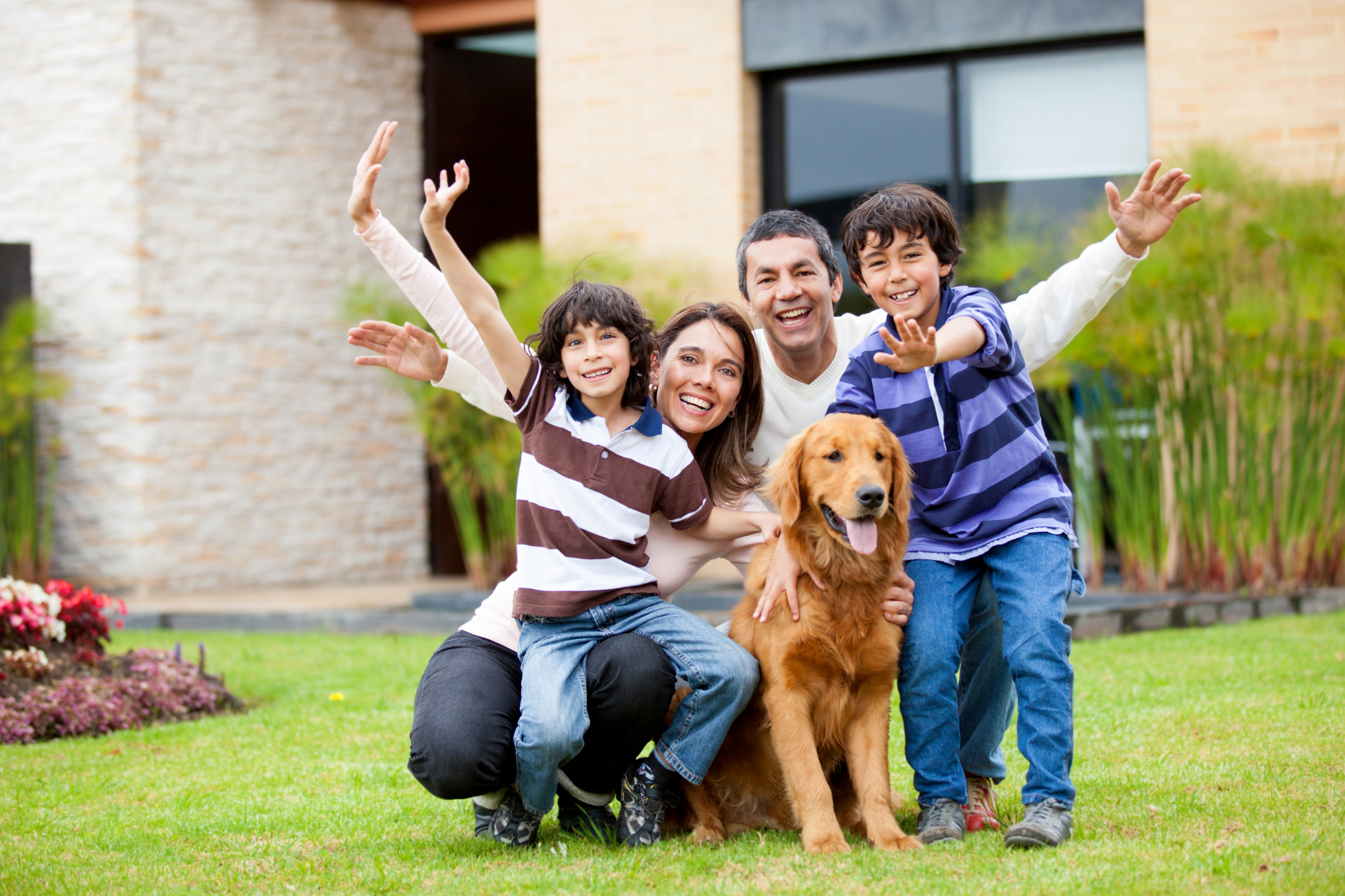 Family posing with a golden retriever on a lawn in front of a house, smiling and waving
