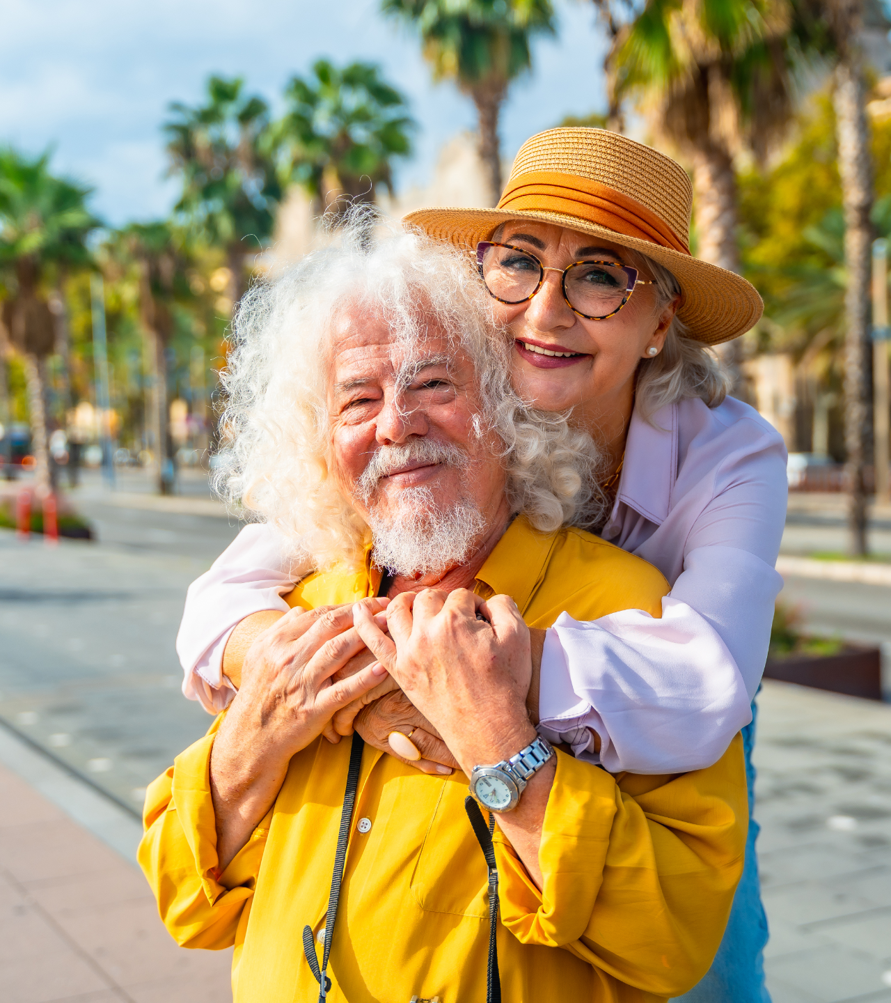 Smiling couple hugging on a sunny palm-lined walkway; woman in straw hat, man in yellow shirt.