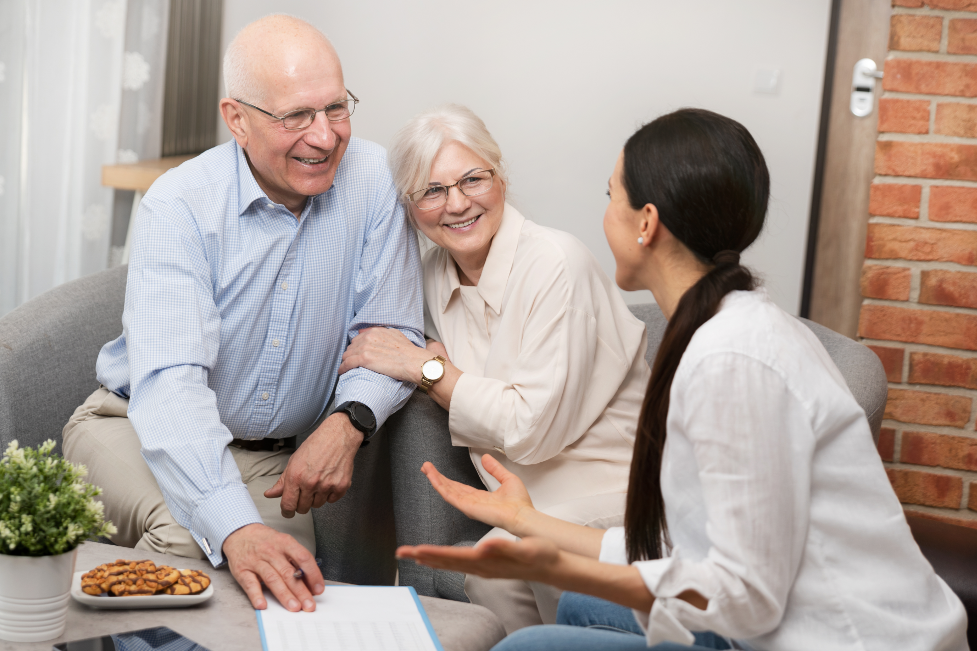 Two people talking with an older couple in a bright living room, seated near a laptop and snacks.