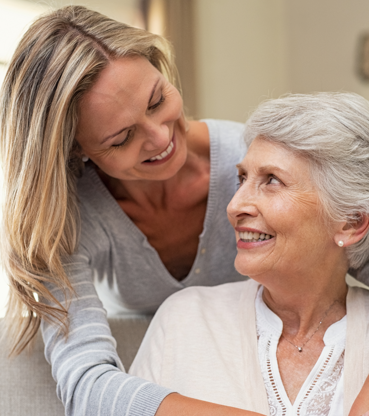 Two smiling women talking indoors, one leaning toward the other in a warm, caring moment