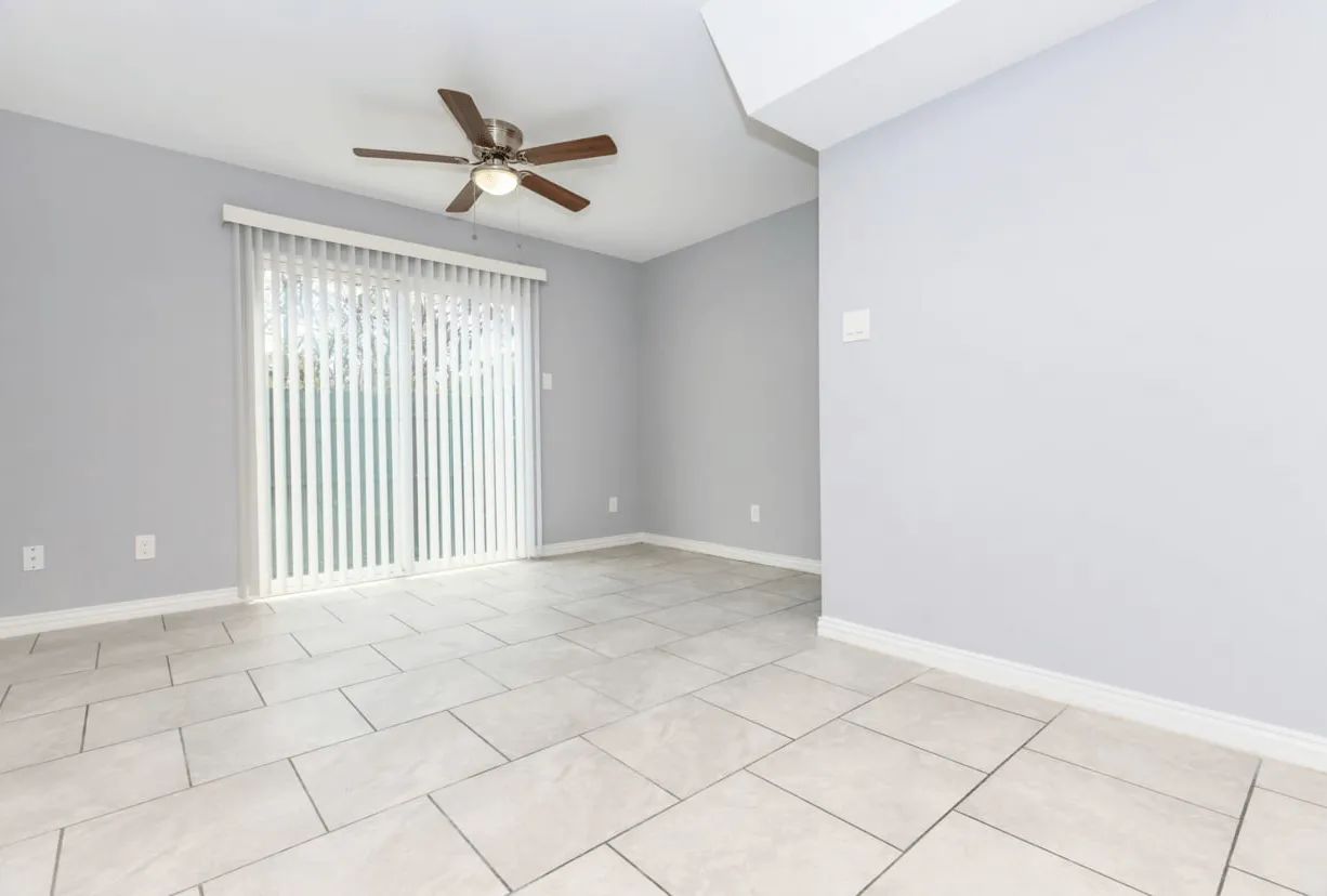 Empty modern living room with light gray walls, tile floor, ceiling fan, and sliding glass door.