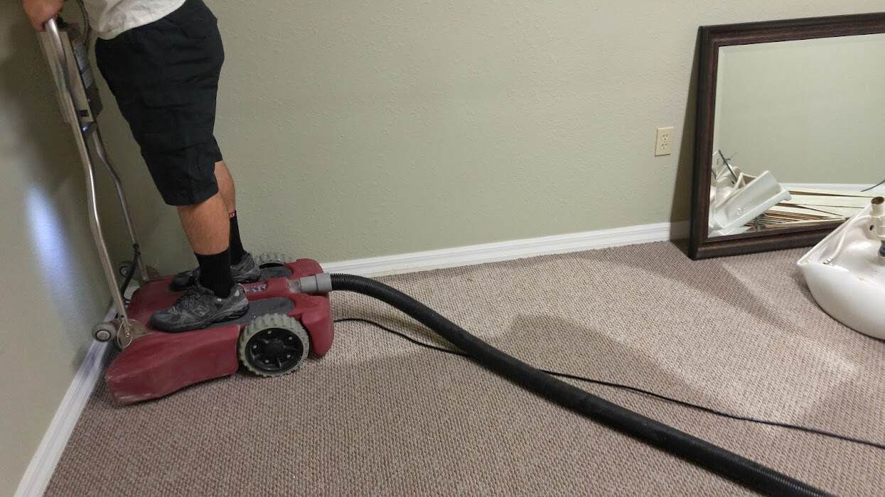 Person using a red floor buffer on beige carpet in a room with a mirror leaning against the wall