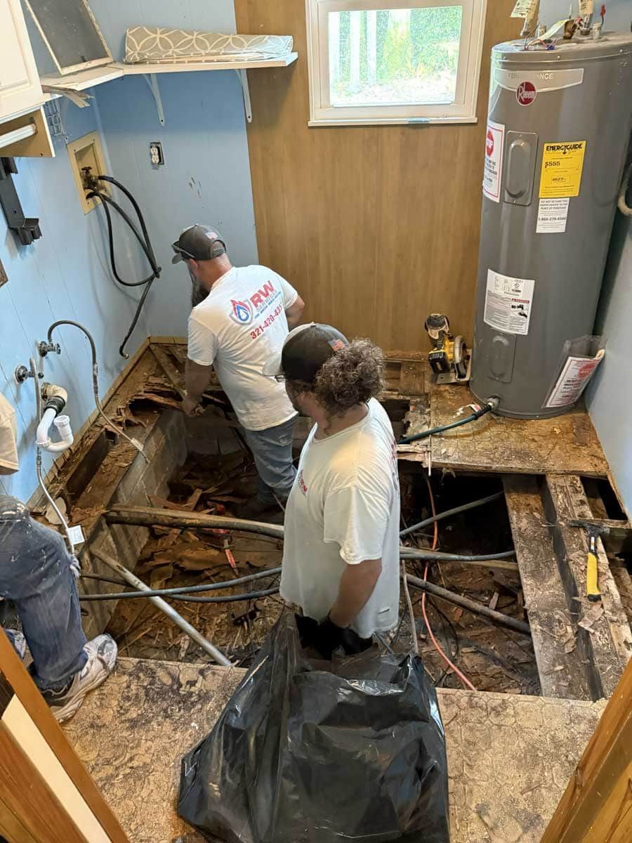 Workers remove damaged flooring in a flooded room with exposed joists and a water heater.