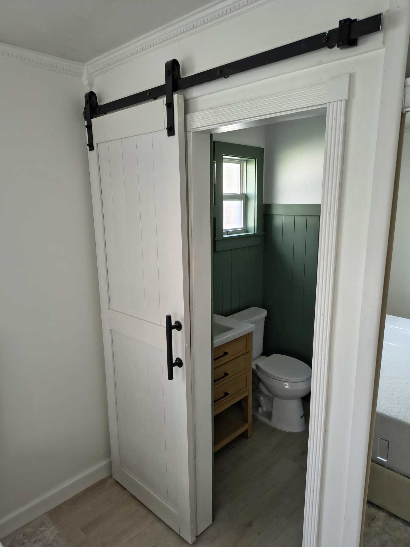 Open barn door revealing a green-and-white bathroom with a toilet and vanity.