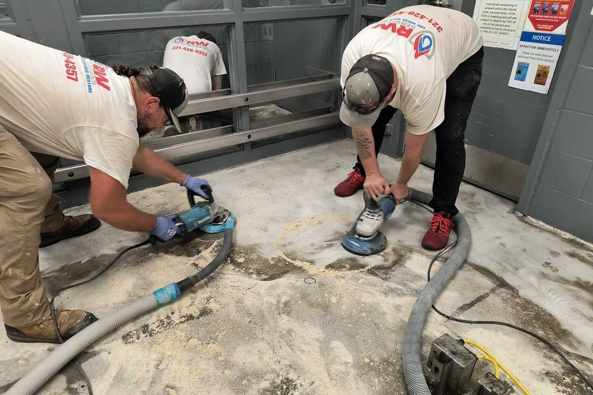 Two workers using power tools to grind a concrete floor in an industrial room