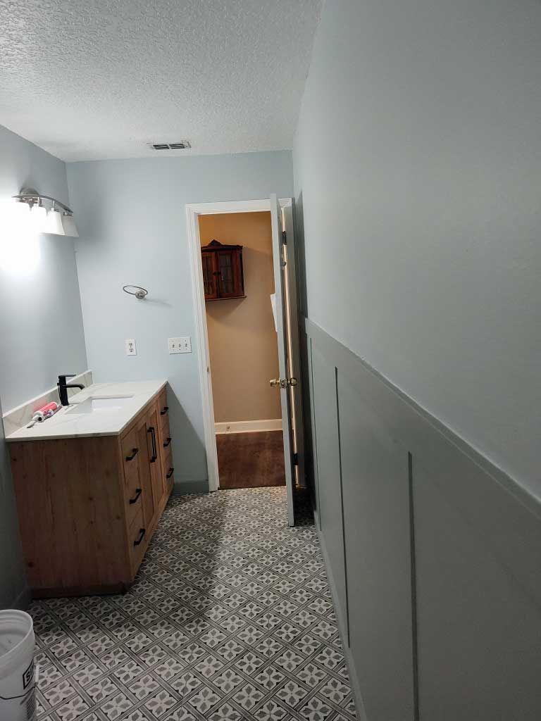 Narrow bathroom with gray wainscoting, patterned tile floor, vanity sink, and open doorway