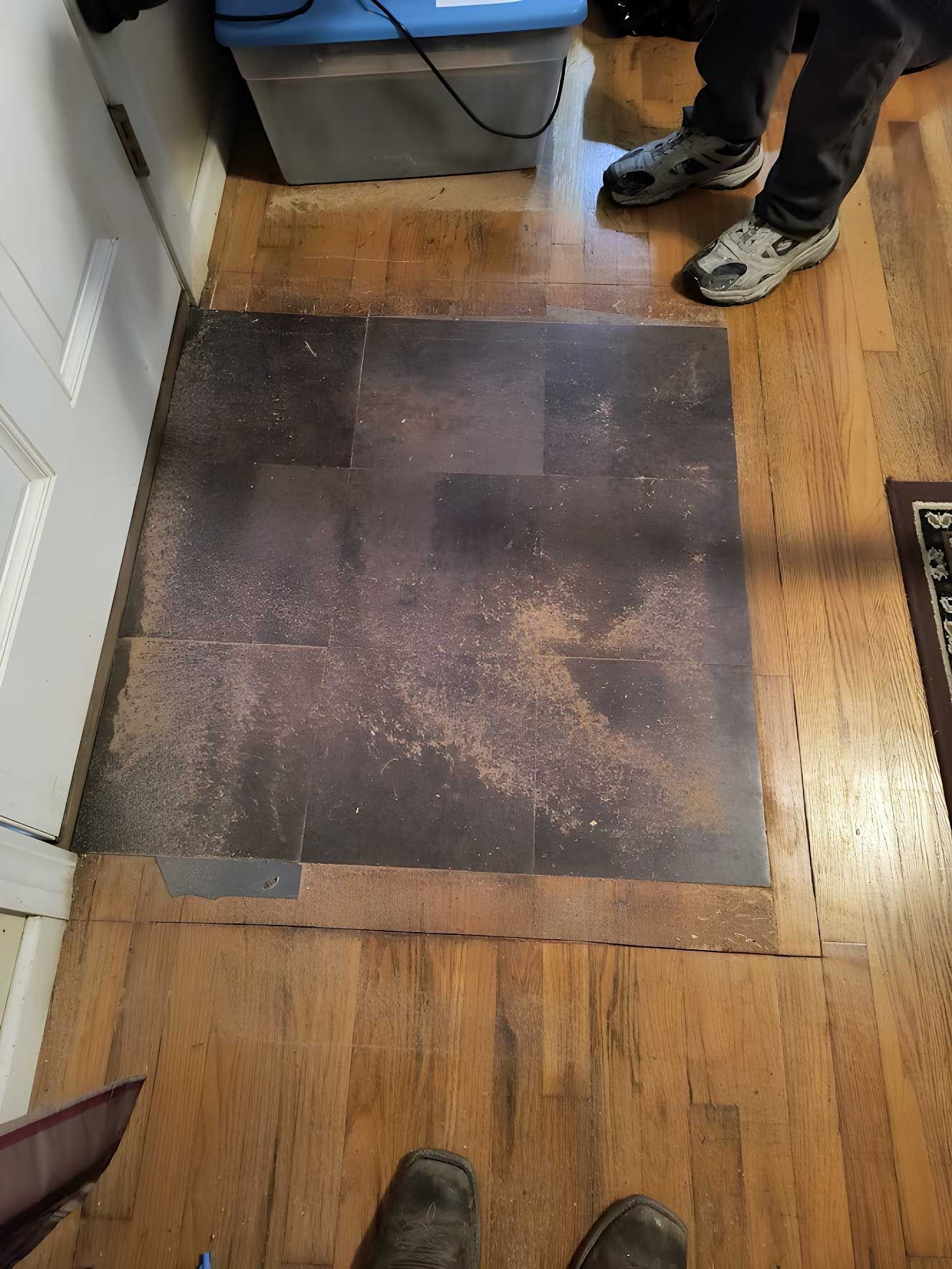 Dark floor mat taped on hardwood near a doorway, with two people’s feet visible at the top and bottom edges.