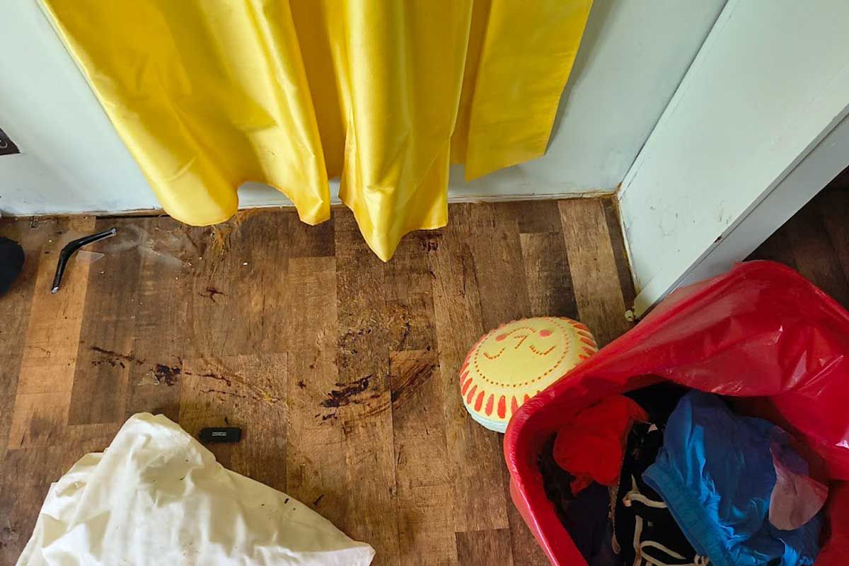Messy floor by yellow curtains with a red bag, white plastic bag, and a small round toy on the right.