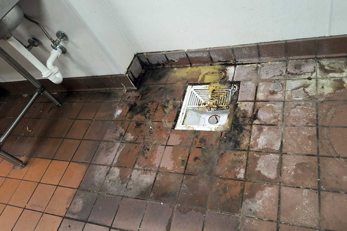 Dirty tiled floor with a small white drain cover and exposed pipes along the wall