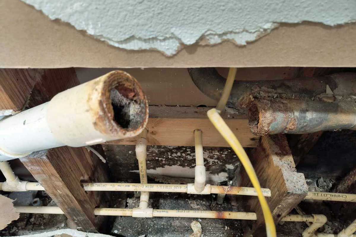 Exposed rusty pipes and damaged ceiling opening with torn drywall and wiring visible