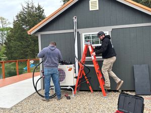 Two technicians install an outdoor HVAC unit on the side of a gray shed, with one standing on a red ladder.