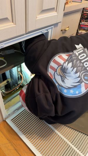 A person wearing a black eagle-print hoodie reaching into an open HVAC return vent located in a cabinet.