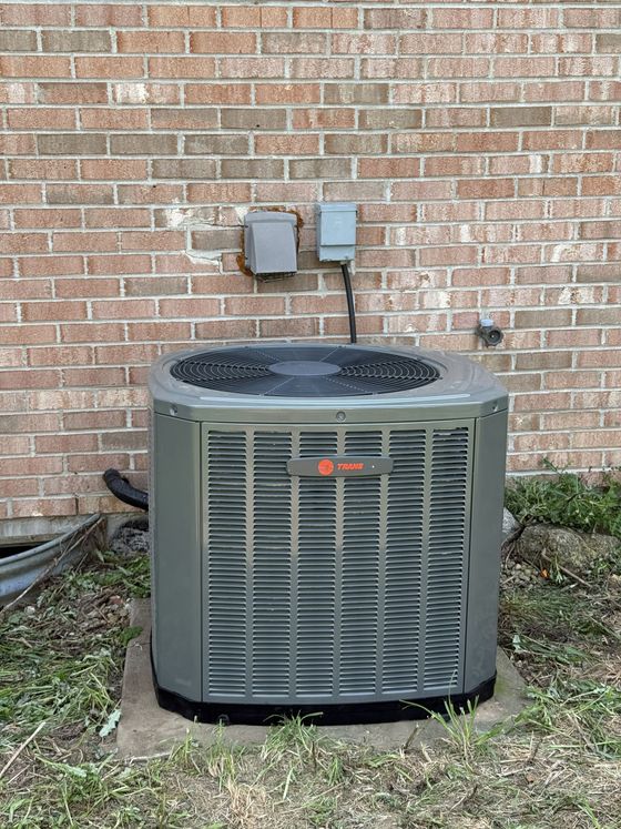 A grey Trane air conditioning unit sits on a concrete pad against a brick wall outside a building.