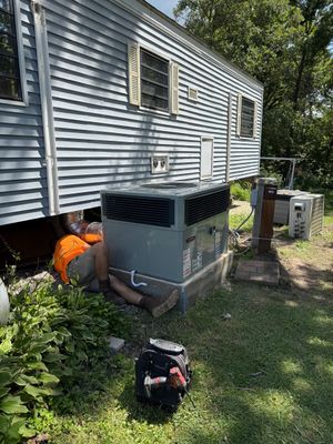 A technician in an orange shirt works on an outdoor air conditioning unit next to a light blue mobile home.