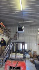 A person in a dark hoodie stands on stairs next to an orange forklift in a warehouse with a wall-mounted heater.