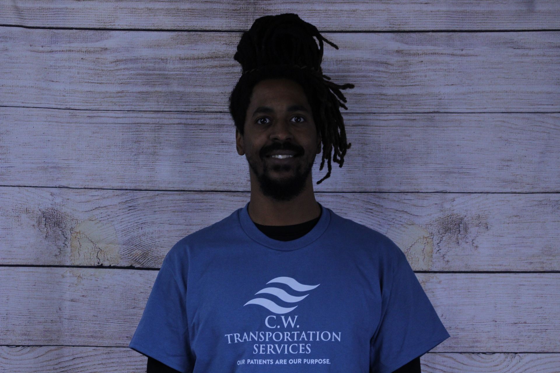 A person with dreadlocks wearing a blue C.W. Transportation Services t-shirt, smiling against a wood-paneled background.
