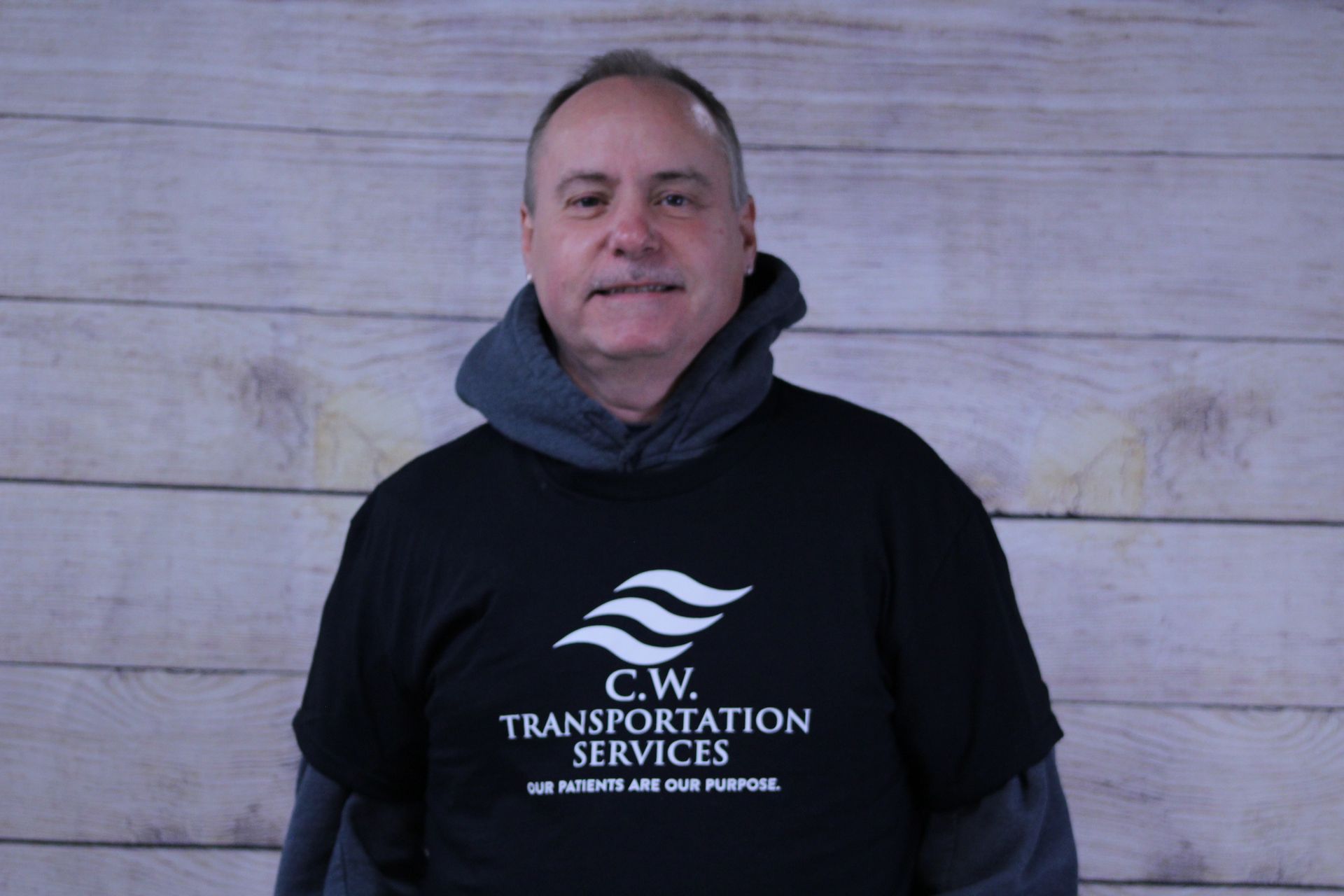 A person wearing a black C.W. Transportation Services t-shirt over a gray hooded sweatshirt against a wood-plank background.