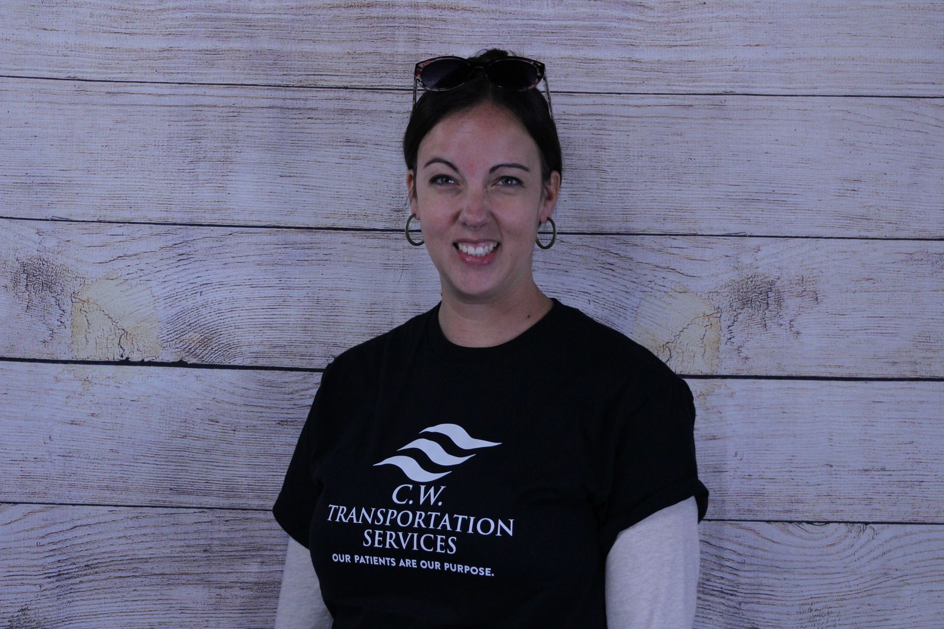 A smiling person wearing a black C.W. Transportation Services t-shirt over a long-sleeved shirt against a wood-paneled wall.