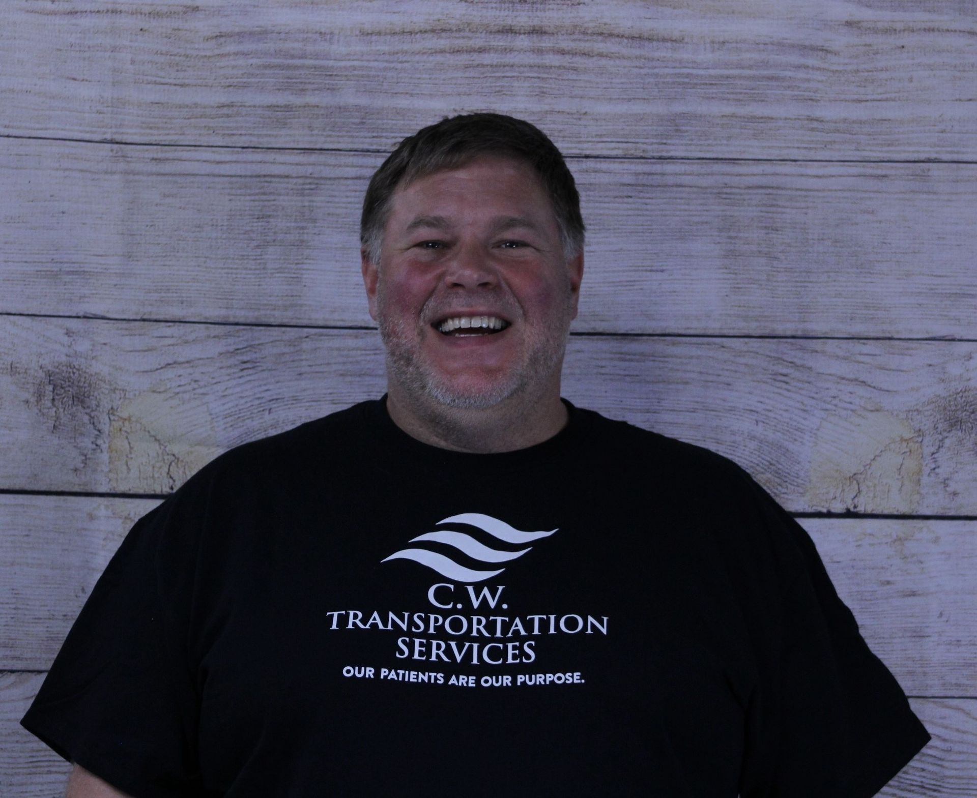 A smiling person wearing a black t-shirt with a white company logo for C.W. Transportation Services against a wood background.